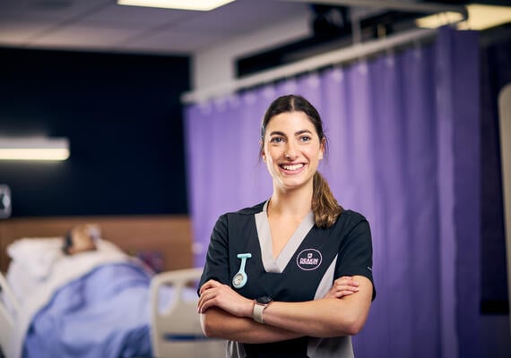 Nursing student standing with crossed arms on a ward and smiling away from camera