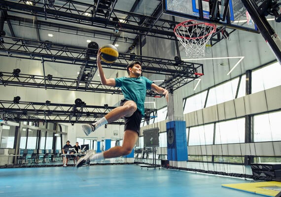 Student about to slam dunk a basketball while two more students watch on from a desk with equipment